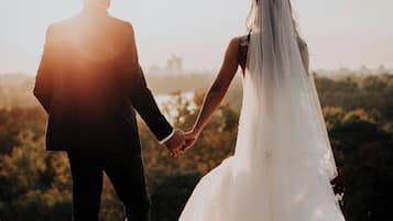 A couple holding hands in a wedding dress and suit, standing on a grassy field with a sunset in the background.