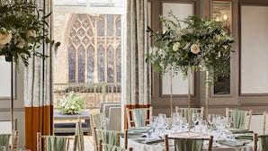 A dining area with a round table set for a meal, surrounded by chairs with green upholstery, and a large floral arrangement above the table.