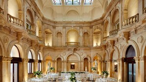 A grand hall with multiple round tables set for dining, ornate columns, and a high ceiling with stained glass windows.