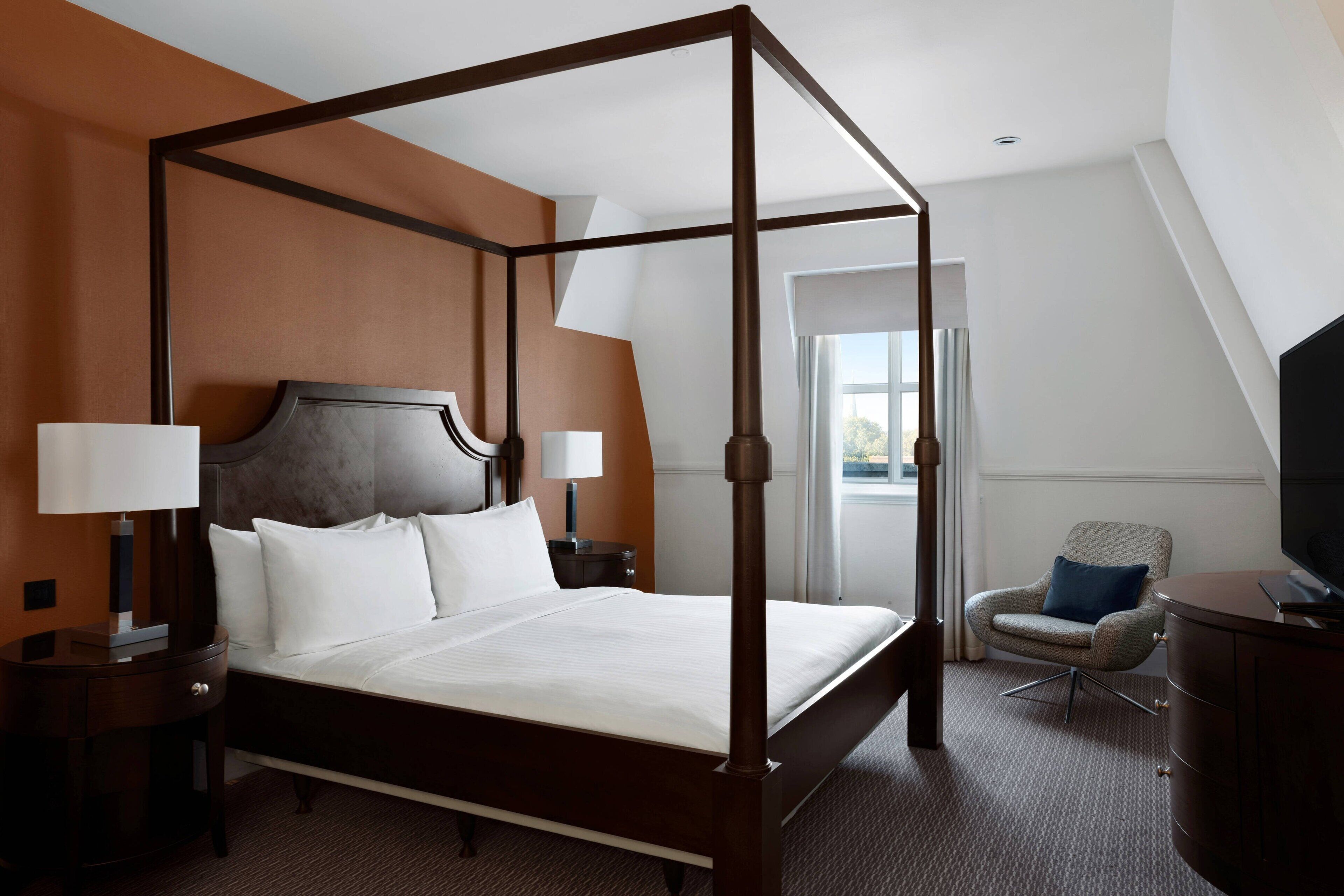 A four-poster bed with a headboard, two bedside tables with lamps, a chair, and a television.