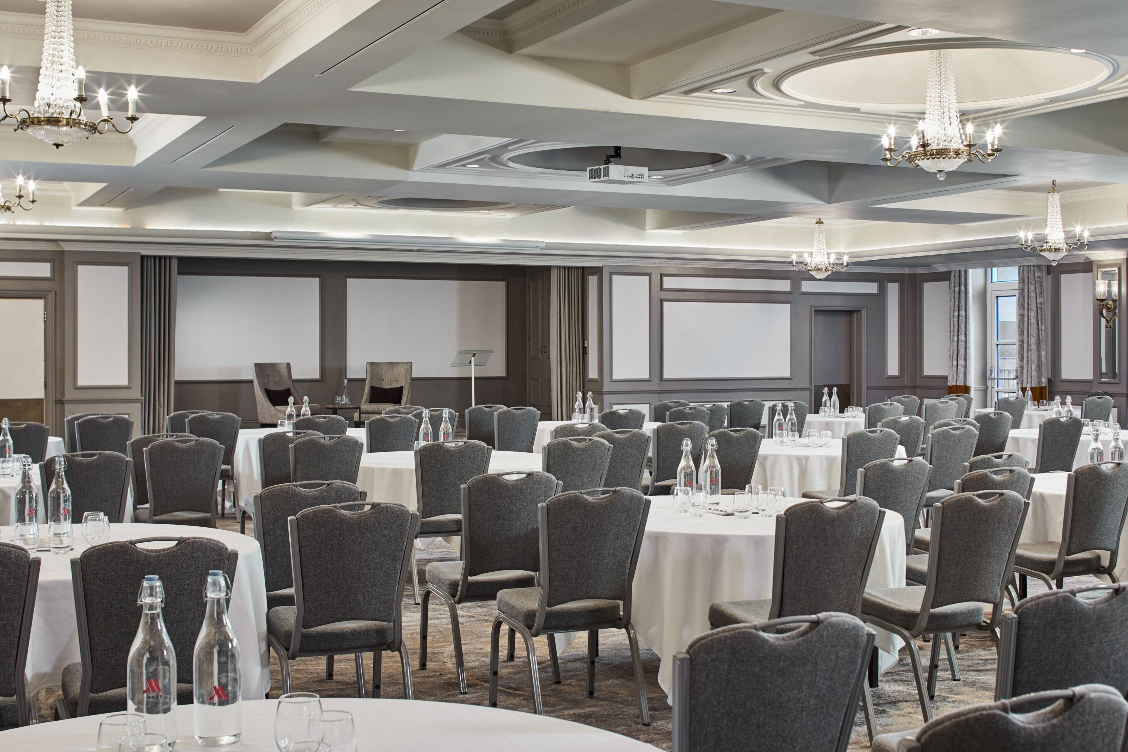 A banquet hall with round tables, grey chairs, and chandeliers.