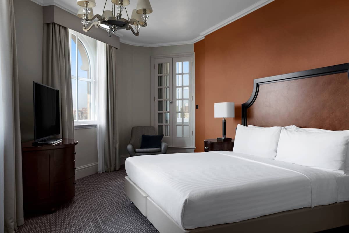 A hotel room with a large bed, a TV on a dresser, a chair, a lamp, and a window with curtains.