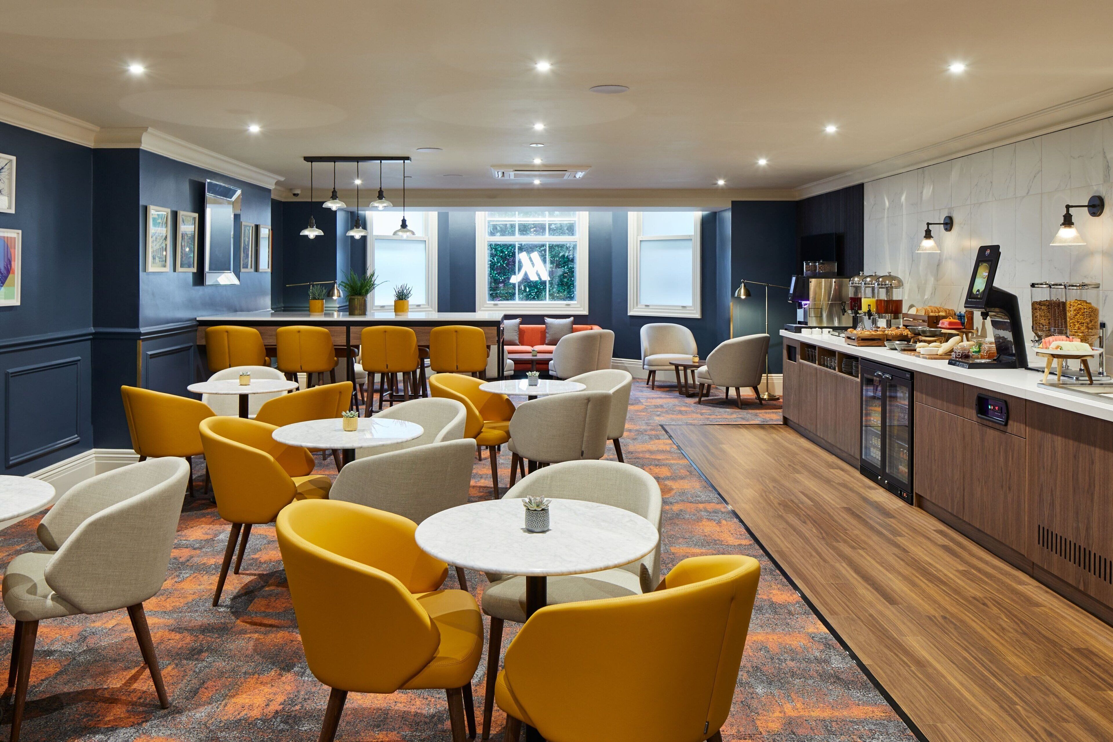Un espace repas moderne avec des chaises jaunes, des tables blanches et un comptoir pour le petit-déjeuner où sont présentés divers aliments.