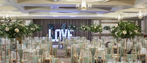 A banquet hall with rows of tables set for a formal event, featuring floral centerpieces and a large illuminated 'LOVE' sign in the background.
