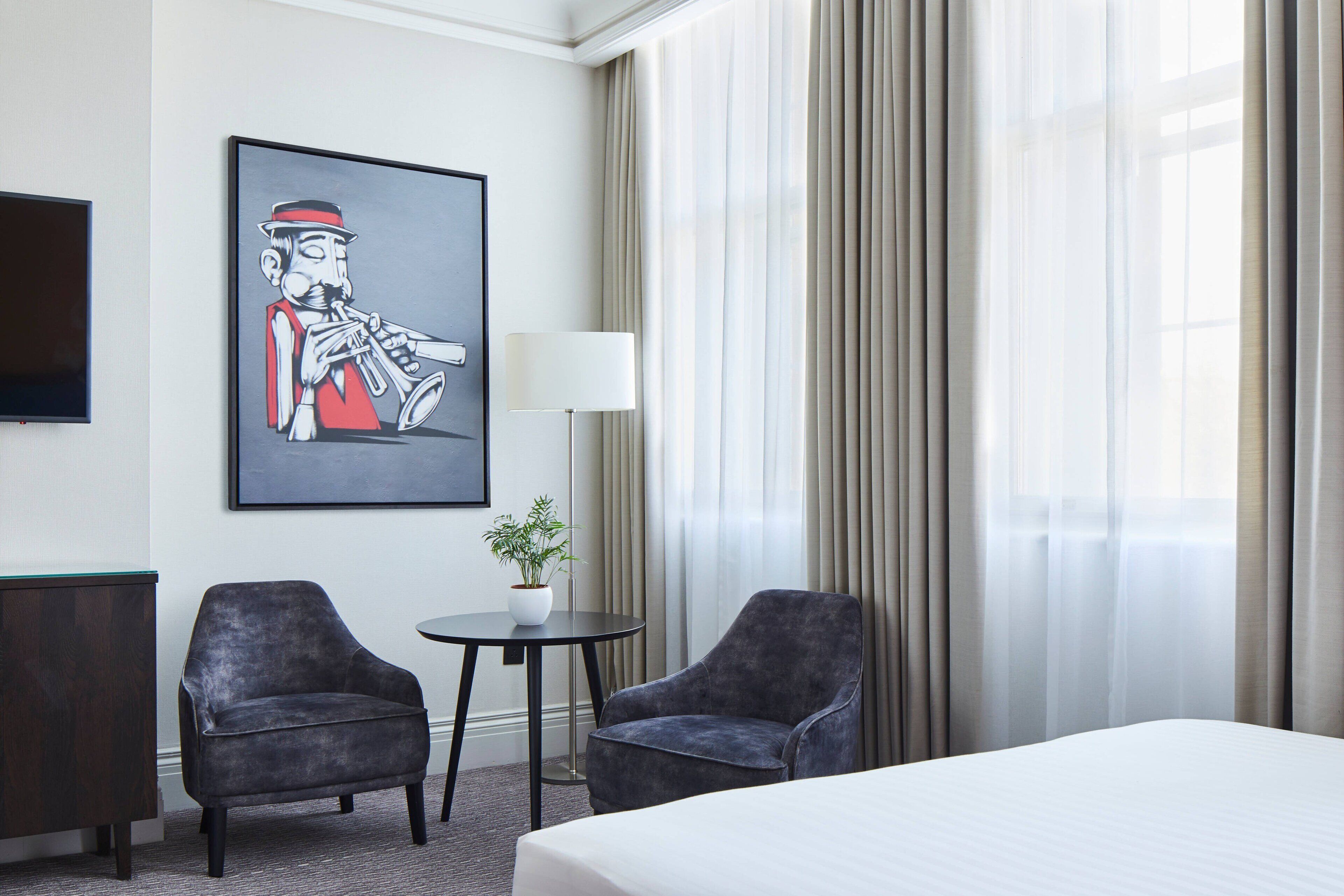 A hotel room with a bed, two armchairs, a small table with a plant, a painting on the wall, and a window with curtains.