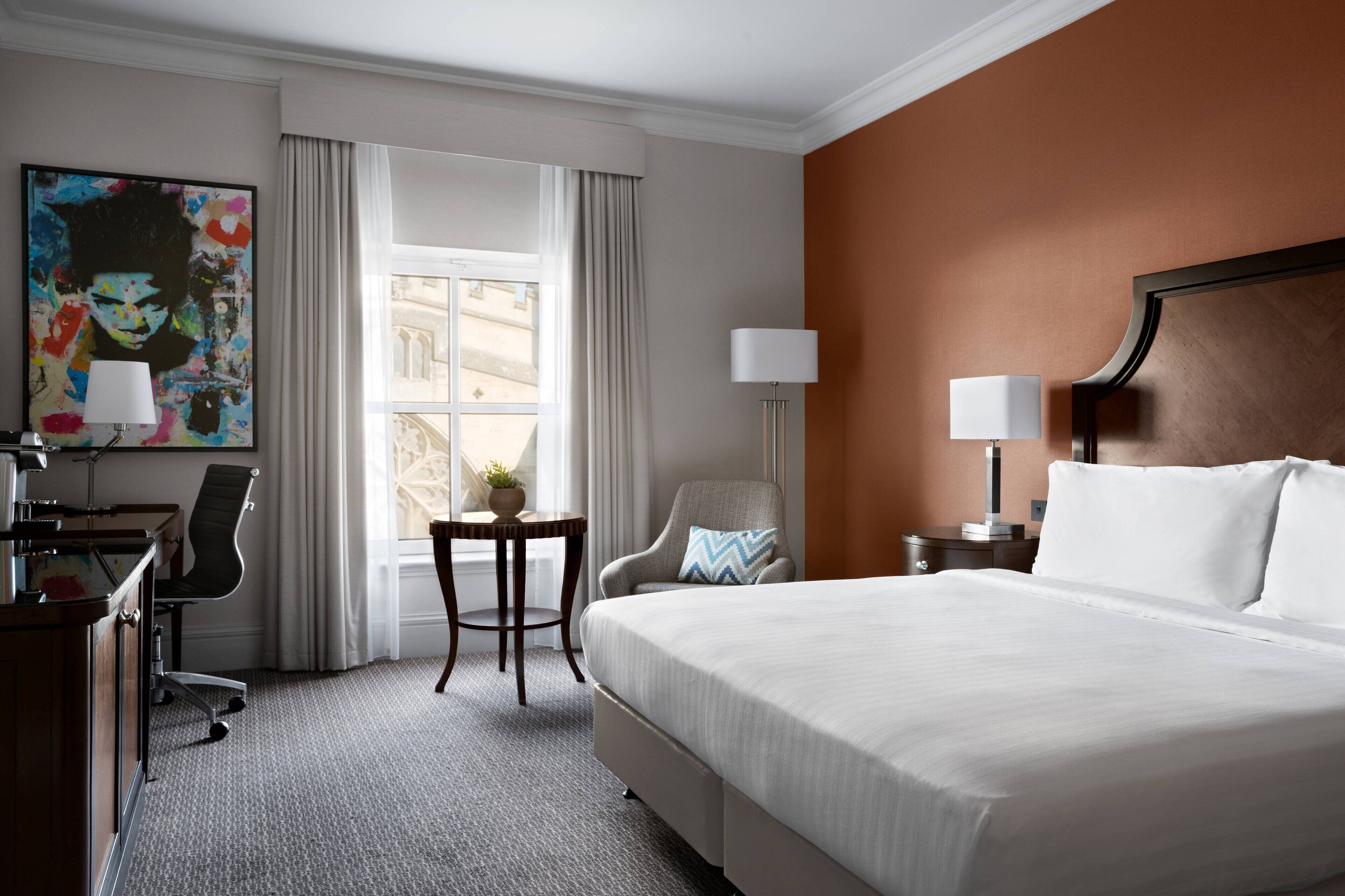 A hotel room with a large bed, a desk with a chair, a small table with a lamp, and a painting on the wall.