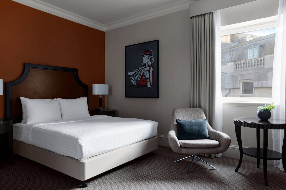 A hotel room with a large bed, a chair, a small table, and a window with a view of buildings.