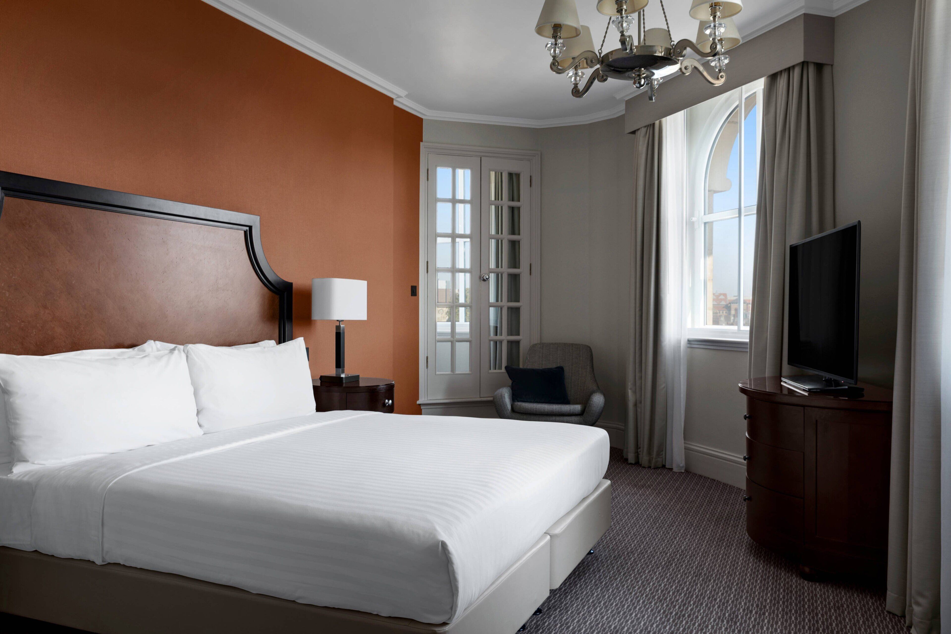 A hotel room with a large bed, a television, a chair, and a desk with a lamp.