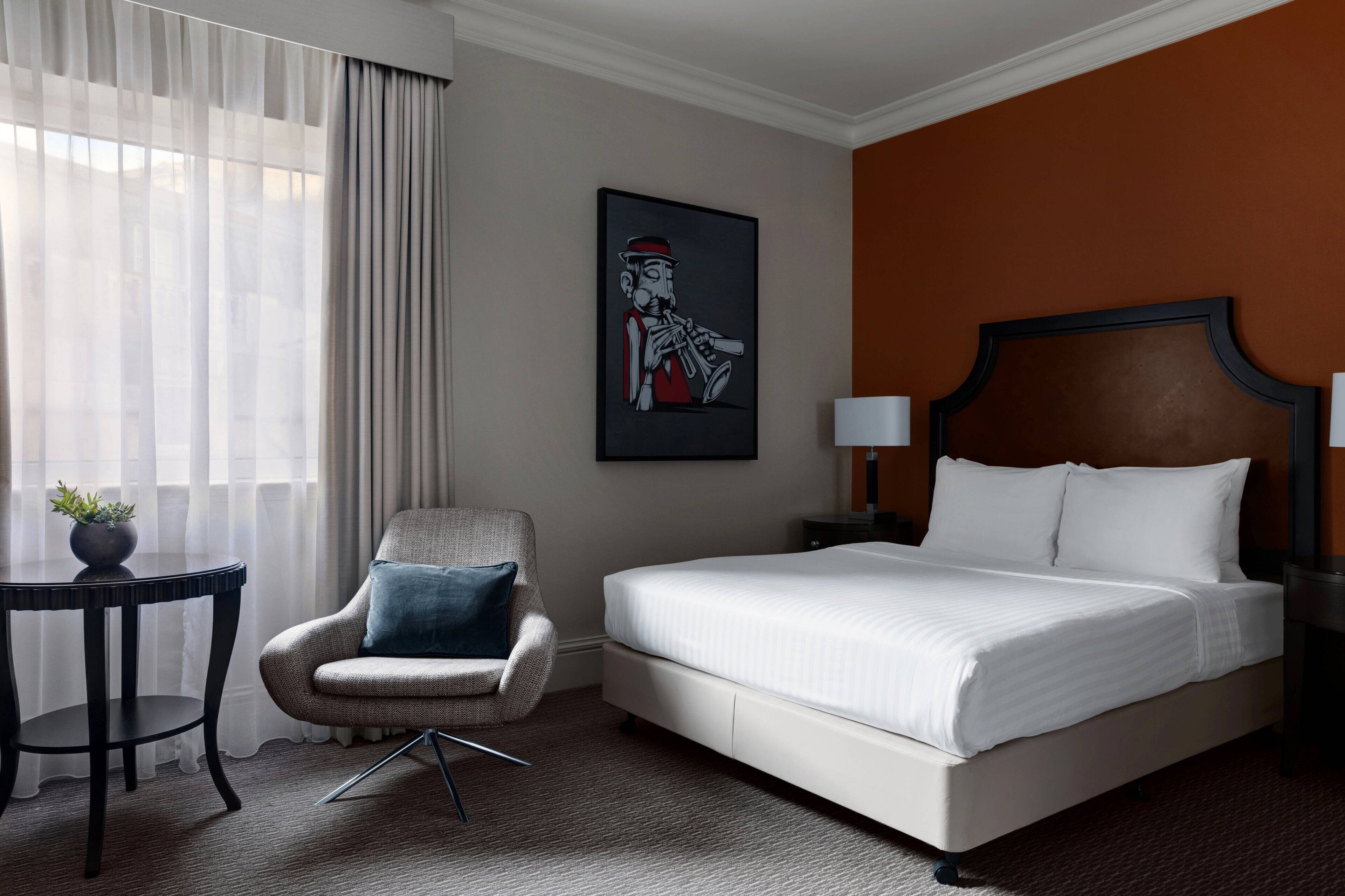 A hotel room with a bed, a chair, a small table with a plant, a painting on the wall, and a nightstand with a lamp.