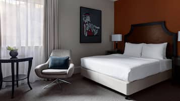 A hotel room with a bed, a chair, a small table with a plant, a painting on the wall, and a nightstand with a lamp.