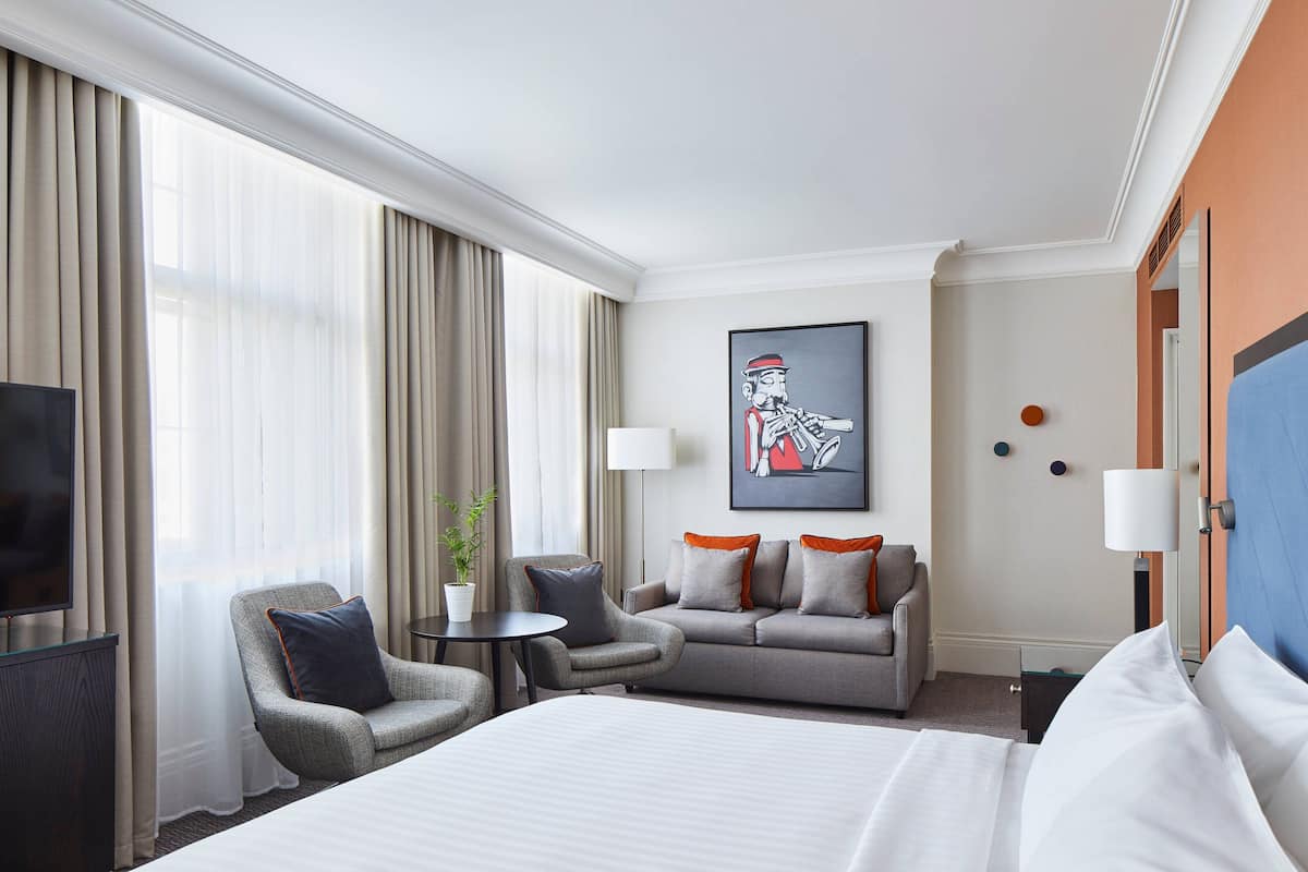 A hotel room with a large bed, a sofa, two armchairs, a TV, and a painting on the wall.