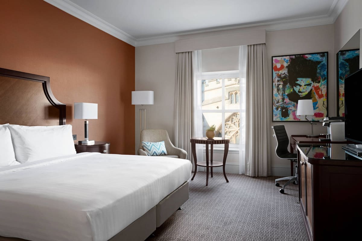 A hotel room with a large bed, a desk with a computer, a chair, a small table with a plant, and a window with curtains.