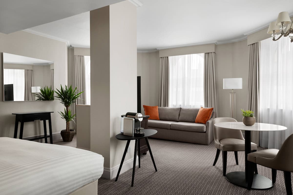 A hotel room with a bed, a sofa, a dining table, and a small side table with a plant.