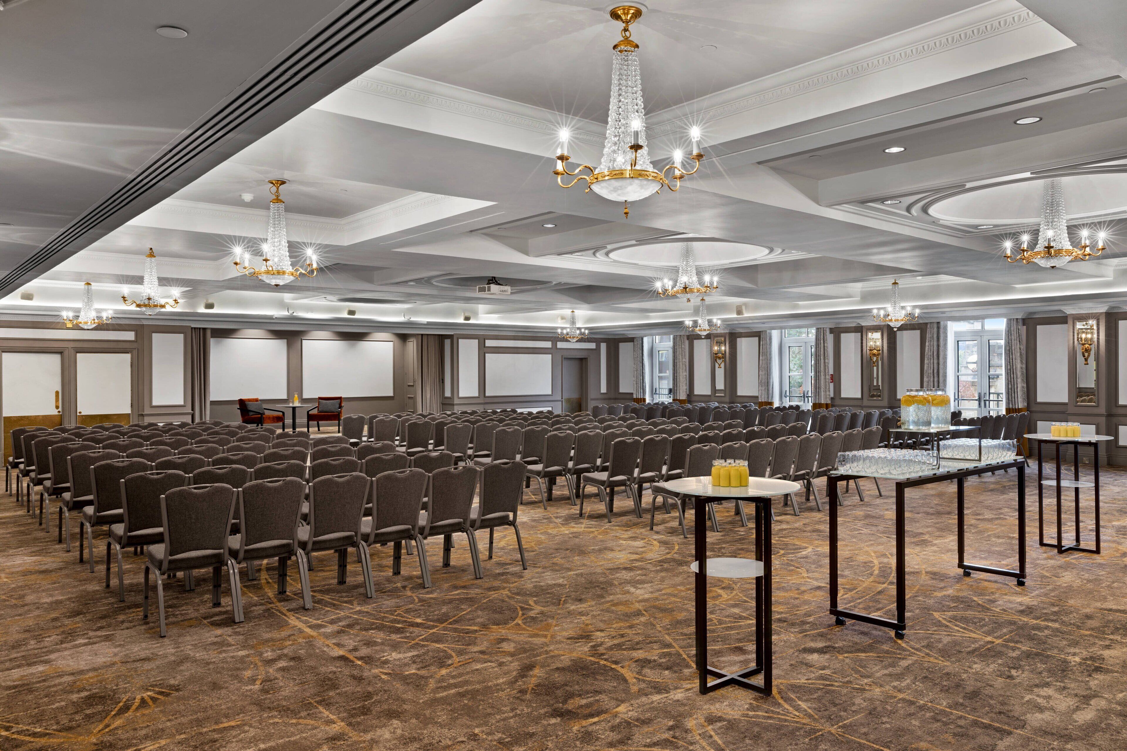 Een ruime vergaderzaal met rijen stoelen, meerdere tafels en decoratieve kroonluchters.