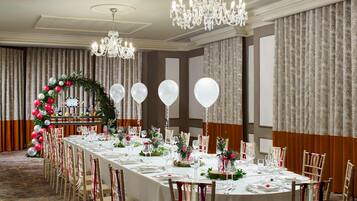Un salón de banquetes con una larga mesa dispuesta para un evento formal, rodeado de sillas adornadas con cintas, un candelabro y un arco decorativo con globos.