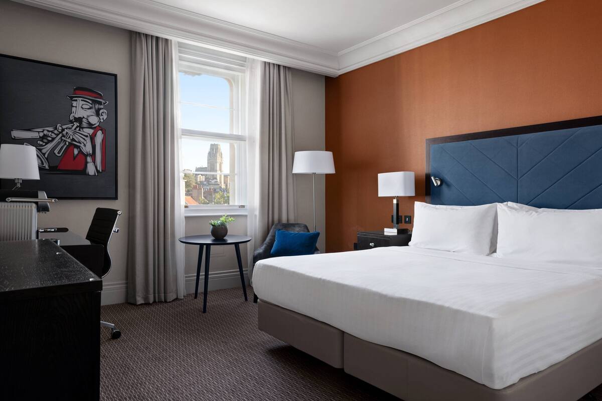 A hotel room with a large bed, a desk, a chair, a small table with a plant, and a painting on the wall.