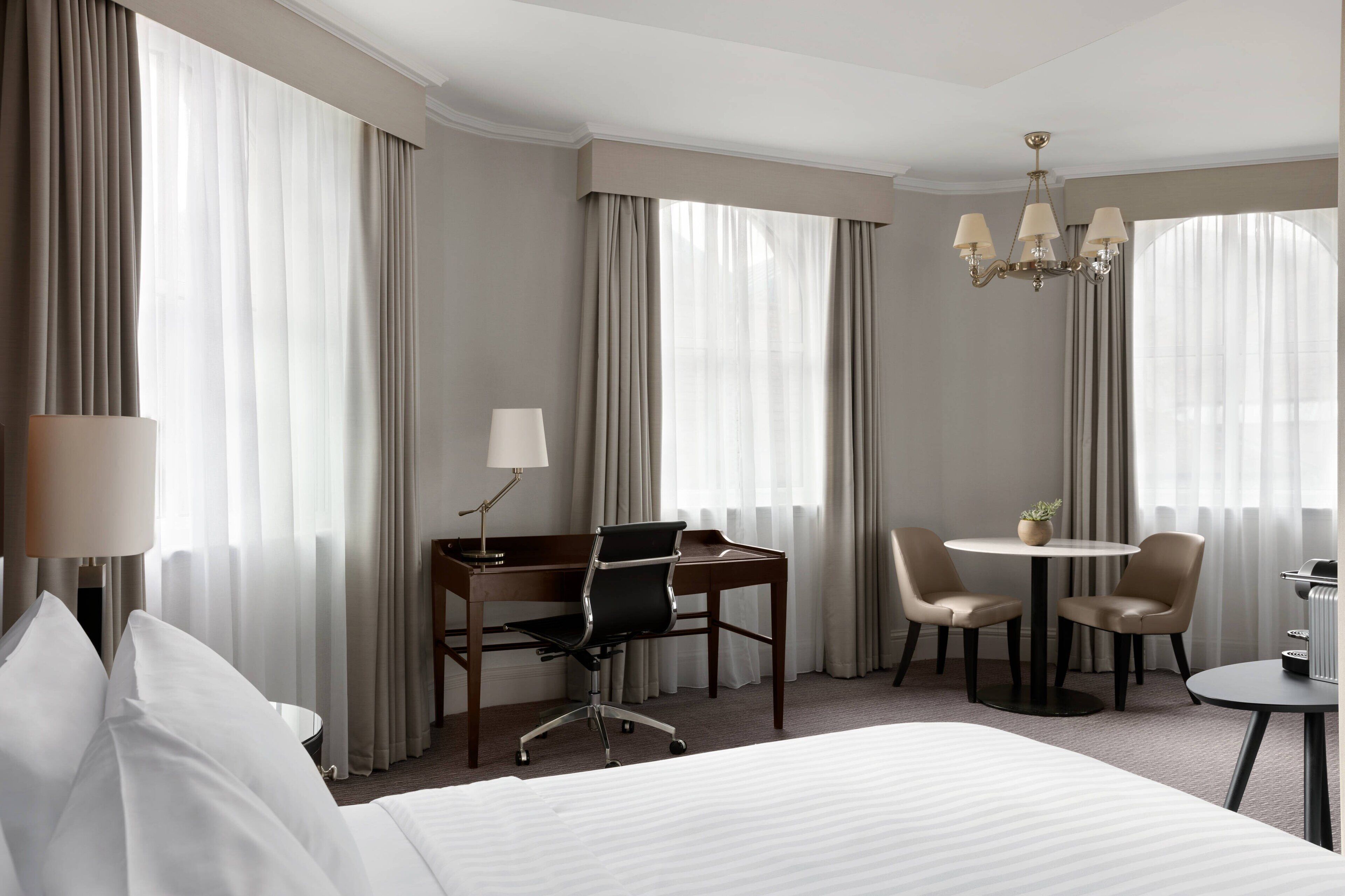 Habitación de hotel con una cama grande, un escritorio con una silla, una mesita redonda con dos sillas y un candelabro.