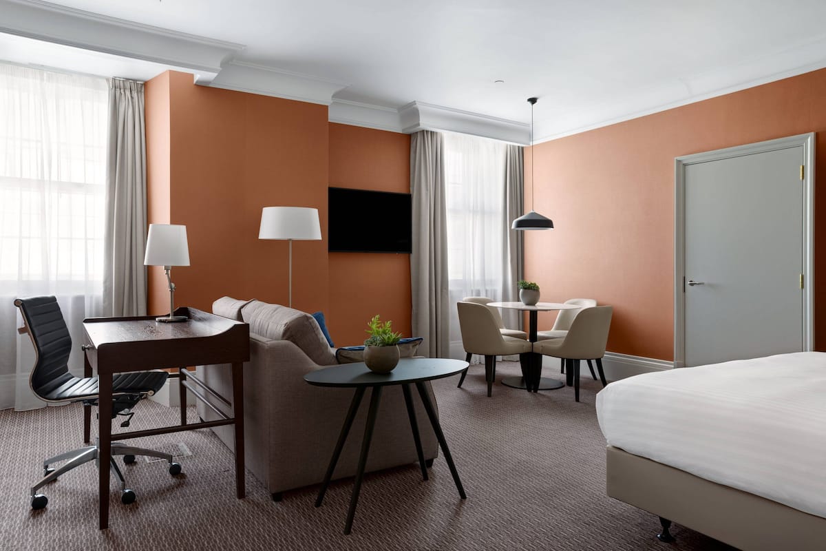 A hotel room with a bed, a desk with a chair, a sofa, a small table with a lamp, and a TV mounted on the wall.