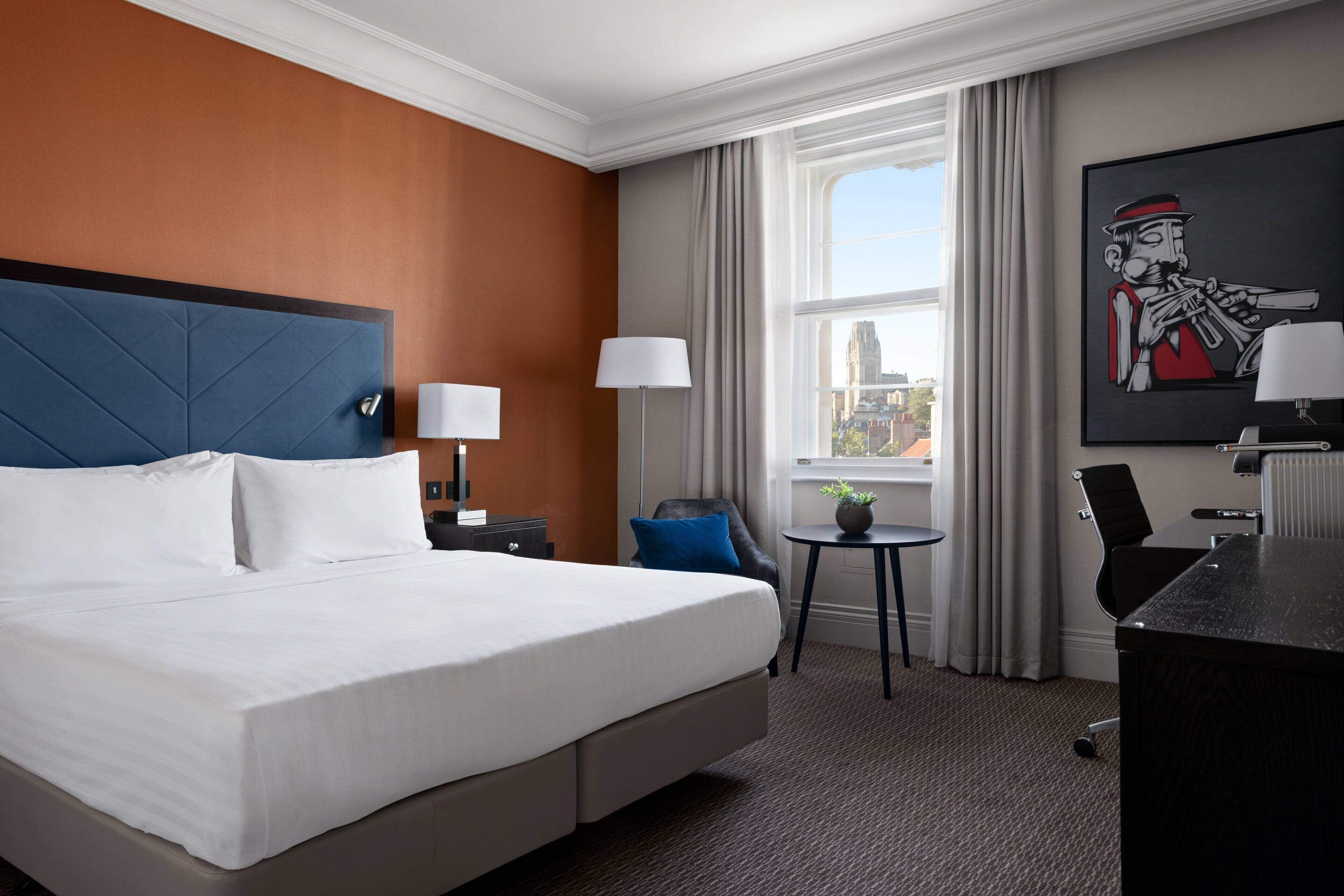 A hotel room with a large bed, a desk, a chair, a small table, and a window with curtains.