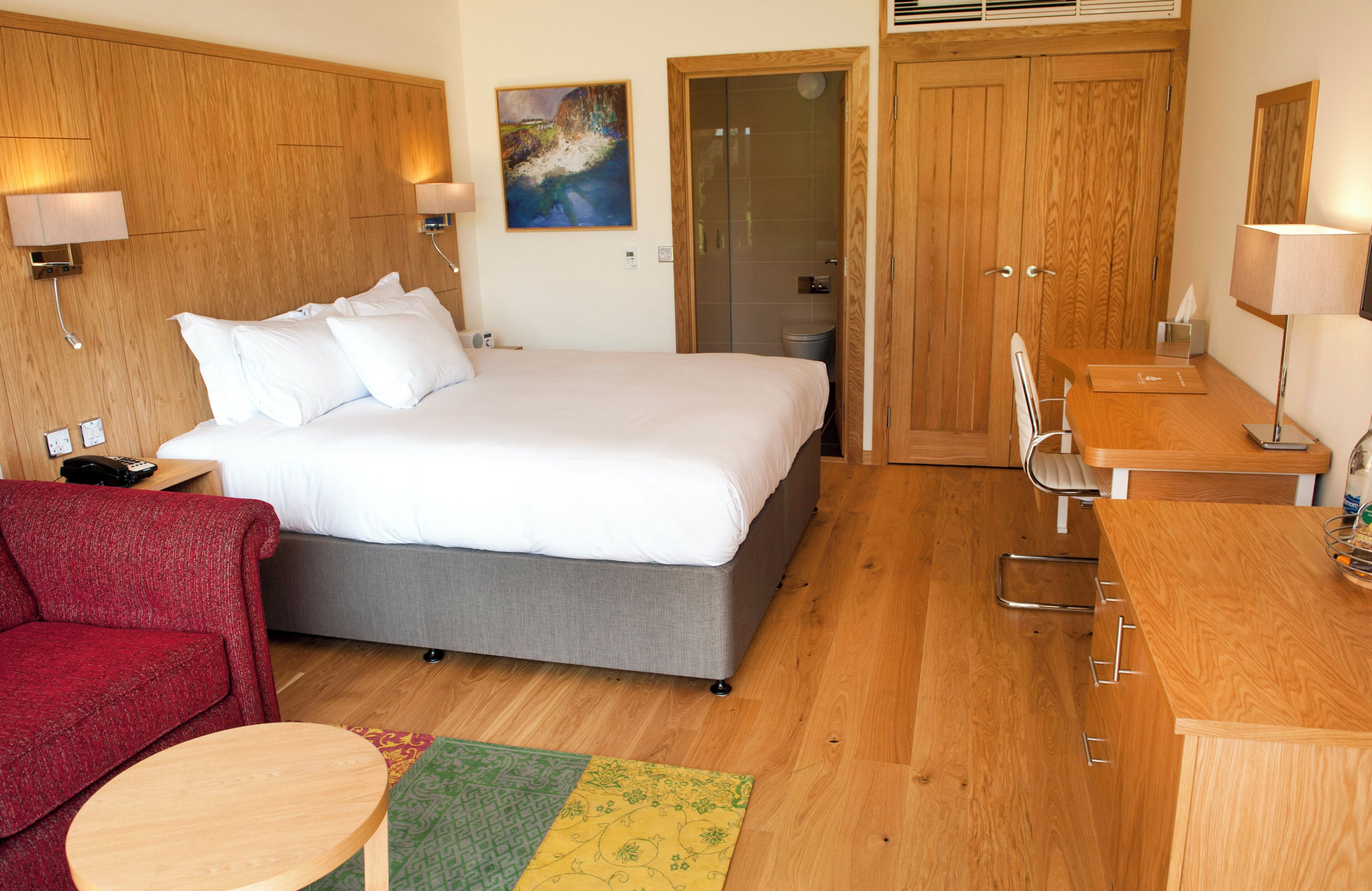 A hotel room with a bed, a desk, a chair, a red sofa, and a small table.