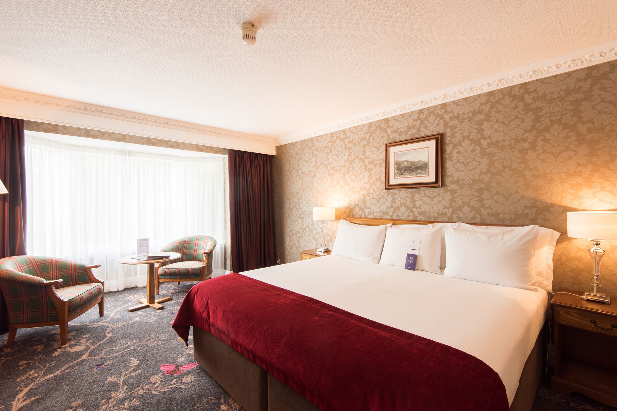 A hotel room with a large bed, two armchairs, a small table, and a nightstand with lamps.