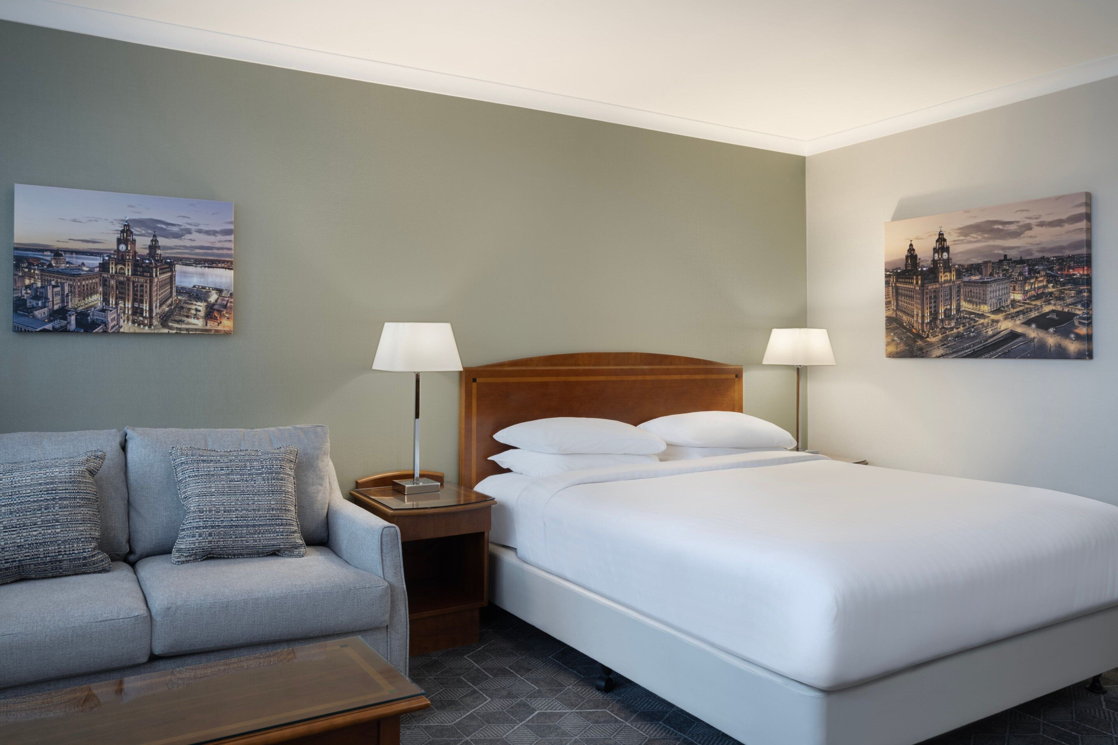 A hotel room with a bed, a sofa, two nightstands with lamps, and two cityscape paintings on the walls.
