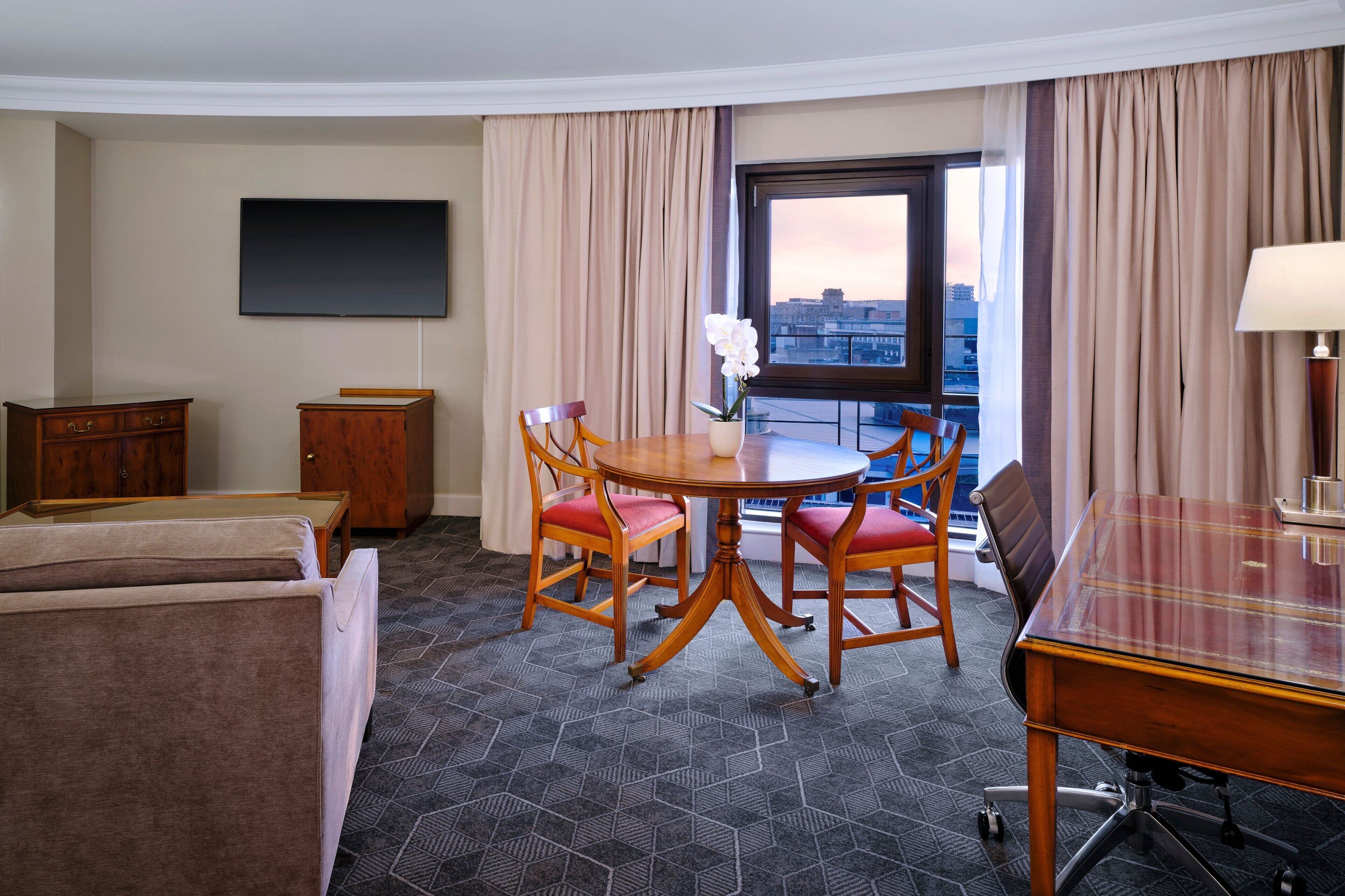 A hotel room with a dining table, chairs, a sofa, a TV, and a window with curtains.
