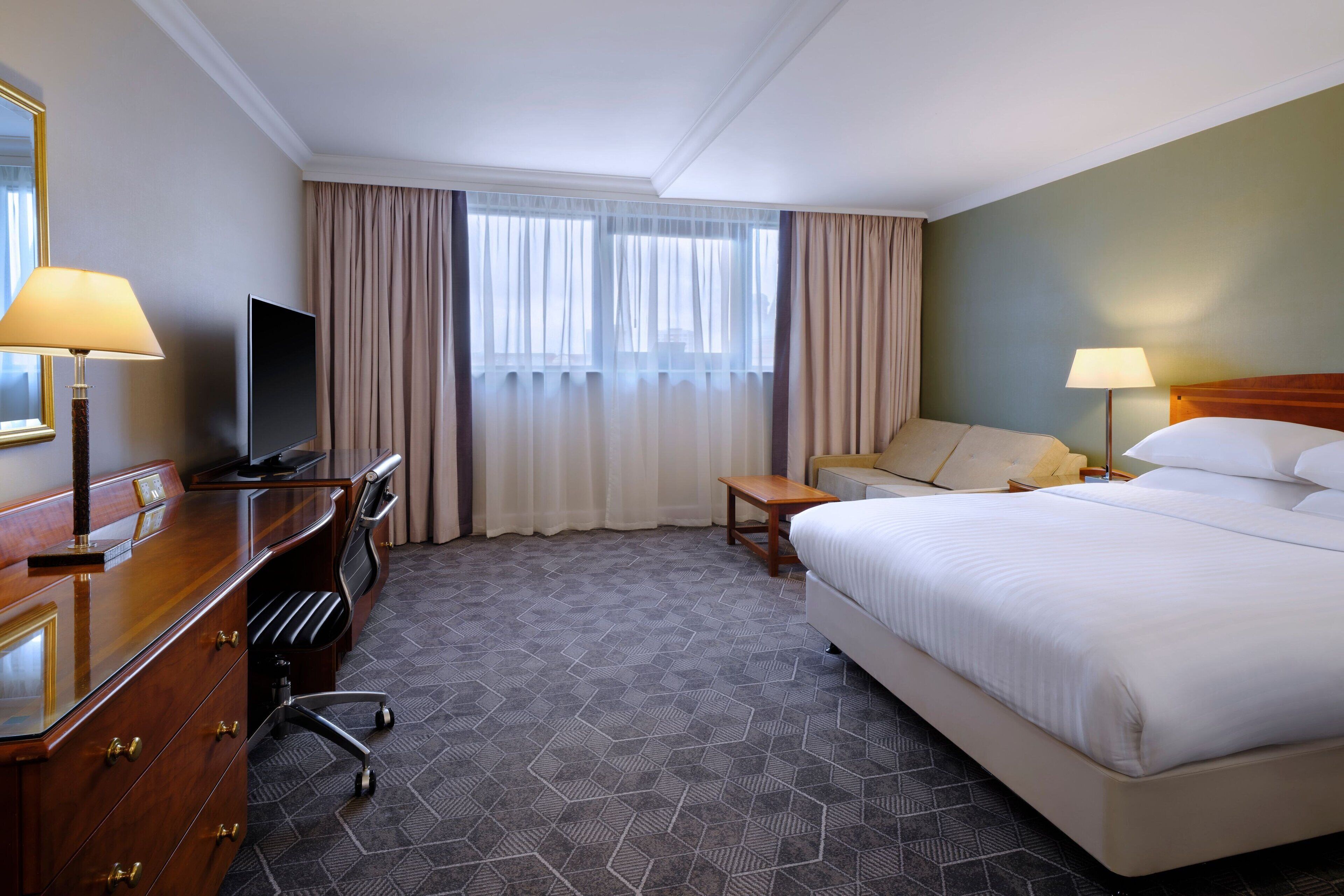 A hotel room with a large bed, a desk with a television, a chair, a small table, and a window with curtains.
