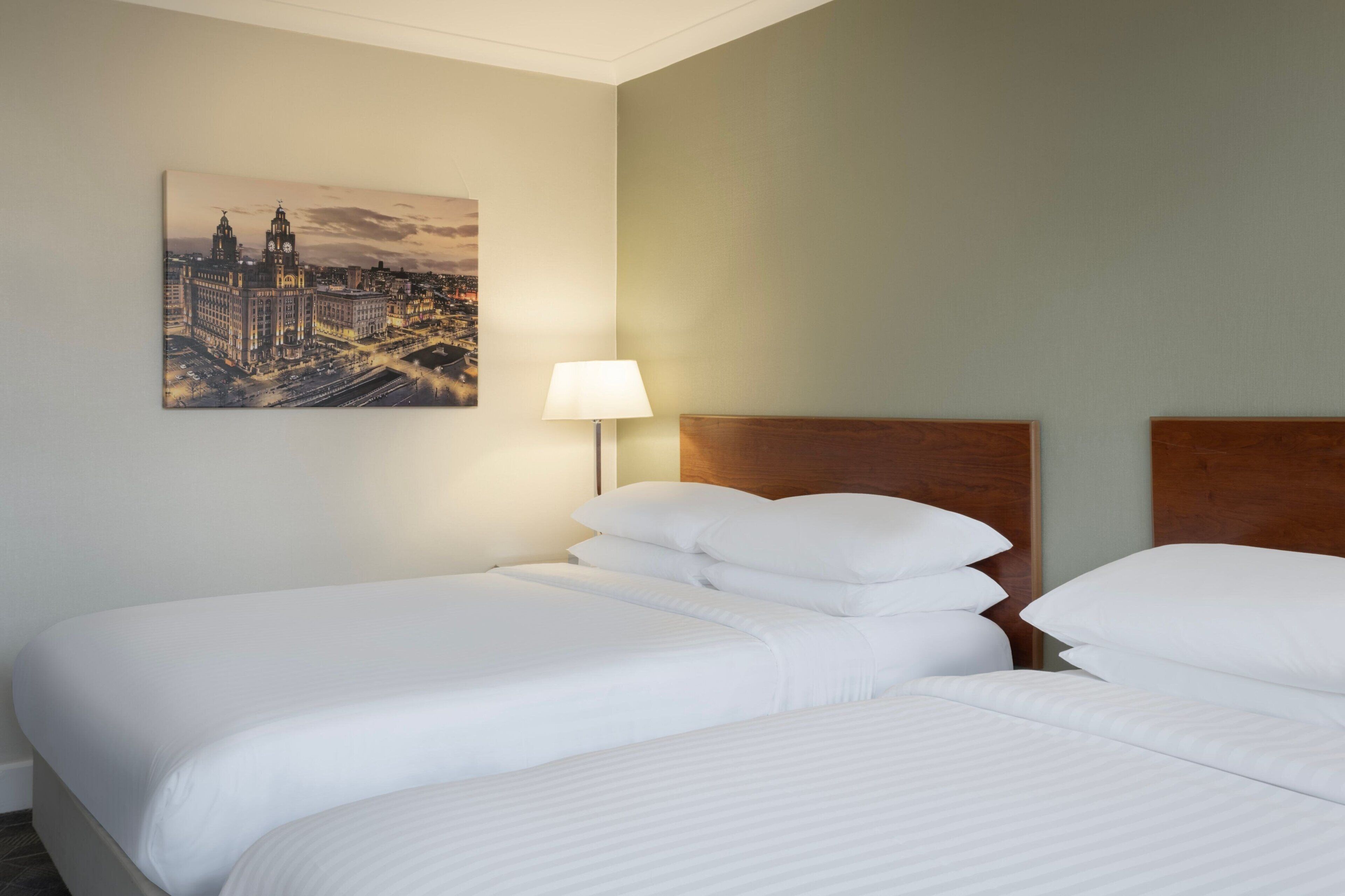 Habitación de hotel con dos camas, una mesita de noche con lámpara y un cuadro de un paisaje urbano en la pared.