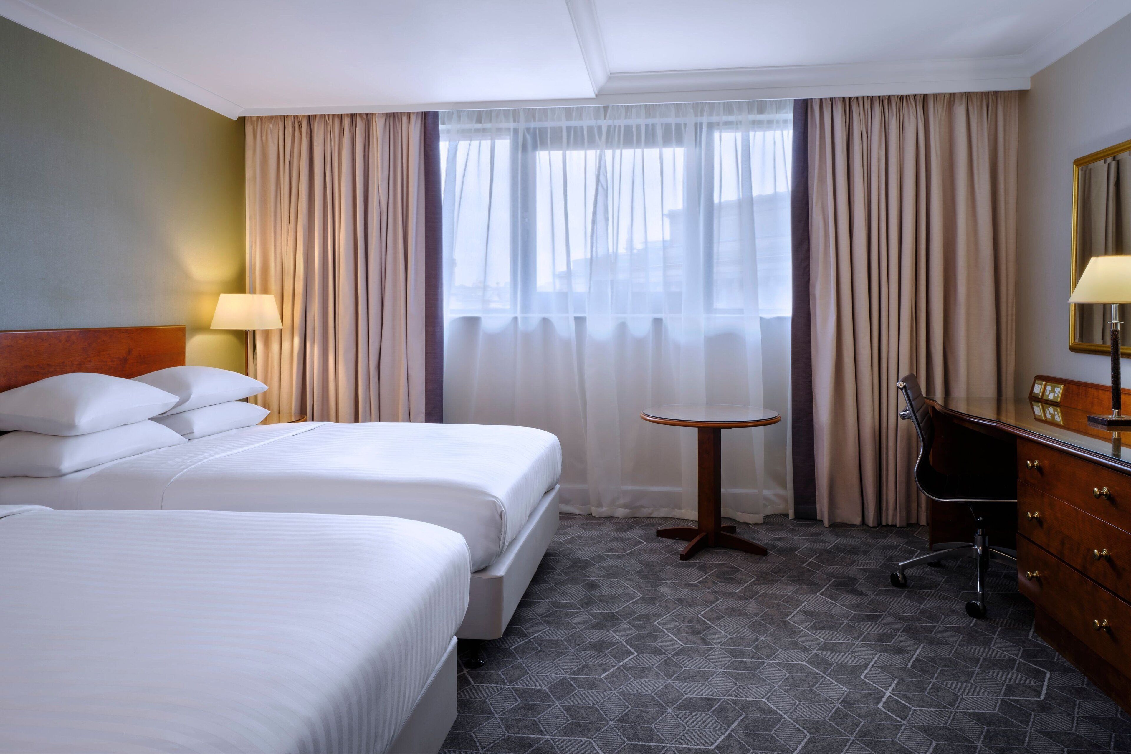 Habitación de hotel con dos camas, una mesa redonda pequeña, un escritorio con espejo y una ventana con cortinas.