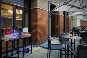 Un espacio de comedor al aire libre con mesas y sillas, una partición de vidrio y un muro de ladrillo.