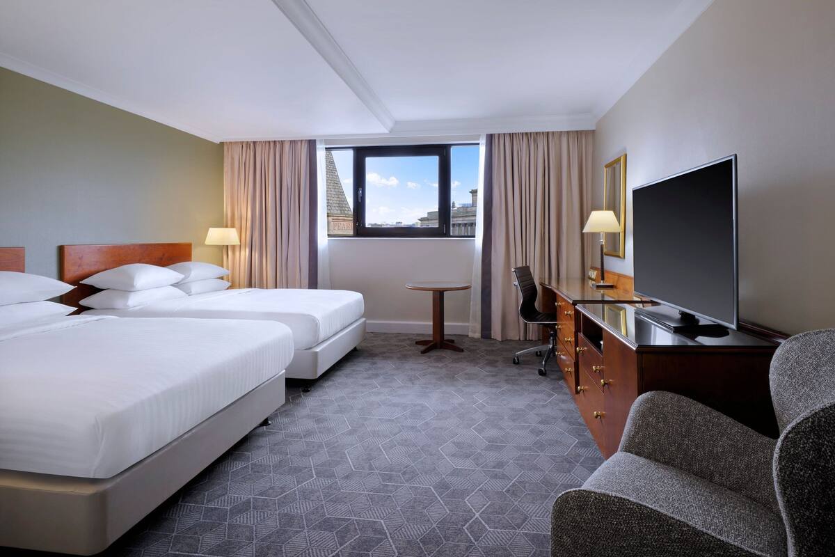 A hotel room with two beds, a desk, a TV, and a view of the city.