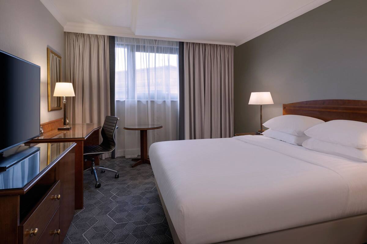 A hotel room with a large bed, a desk with a chair, a television, and a window with curtains.