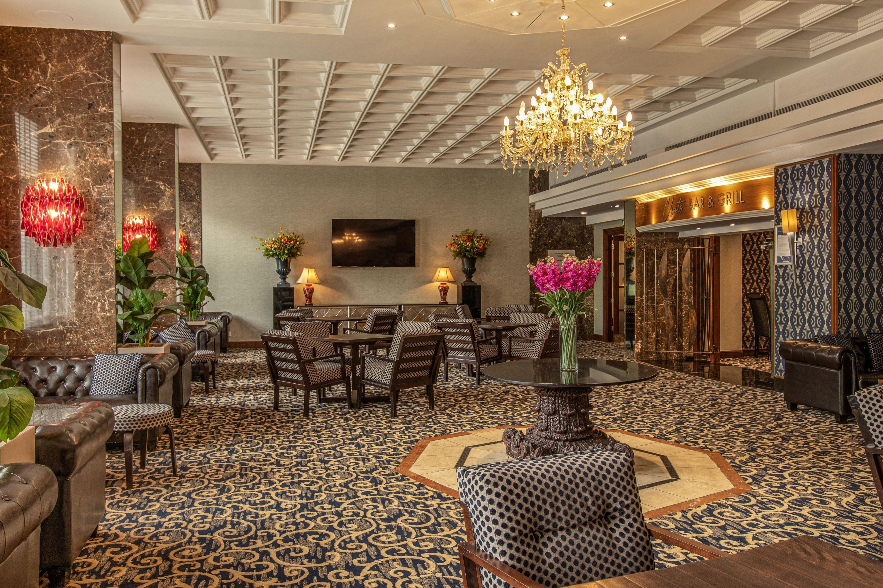 A hotel lobby with a patterned carpet, seating area with sofas and chairs, a chandelier, and a bar area.
