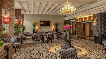 A hotel lobby with a patterned carpet, seating area with sofas and chairs, a chandelier, and a bar area.