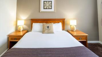 Habitación de hotel con una cama grande, dos mesitas de noche con lámparas, una obra de arte enmarcada sobre la cama y suelo alfombrado.