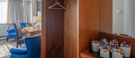 A hotel room with a wooden wardrobe, a desk with a chair, a hanging coat rack, and a coffee station with mugs and cups.