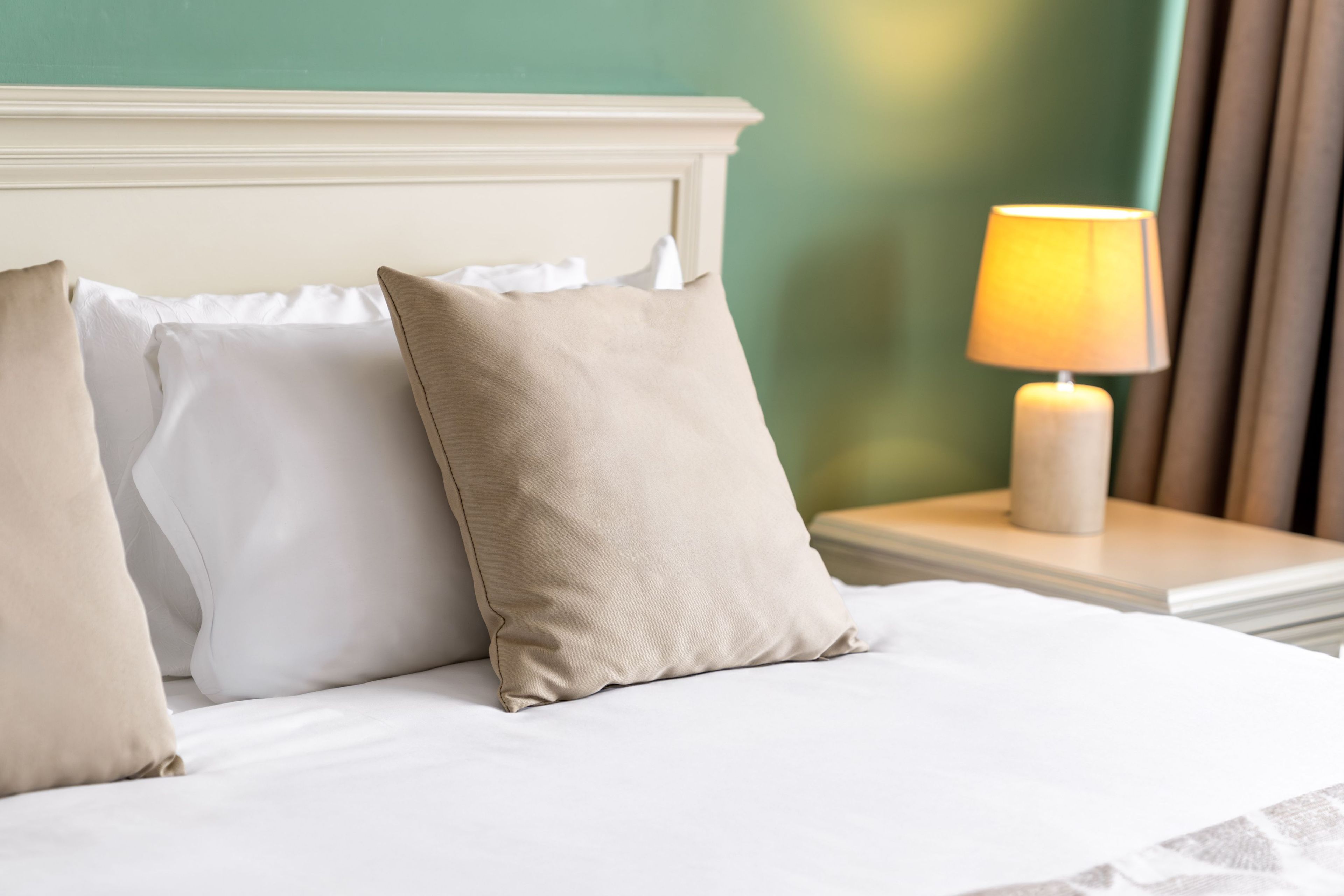 Una cama bien tendida con almohadas blancas y beige, un cabecero blanco y una mesita de noche con una lámpara encendida.