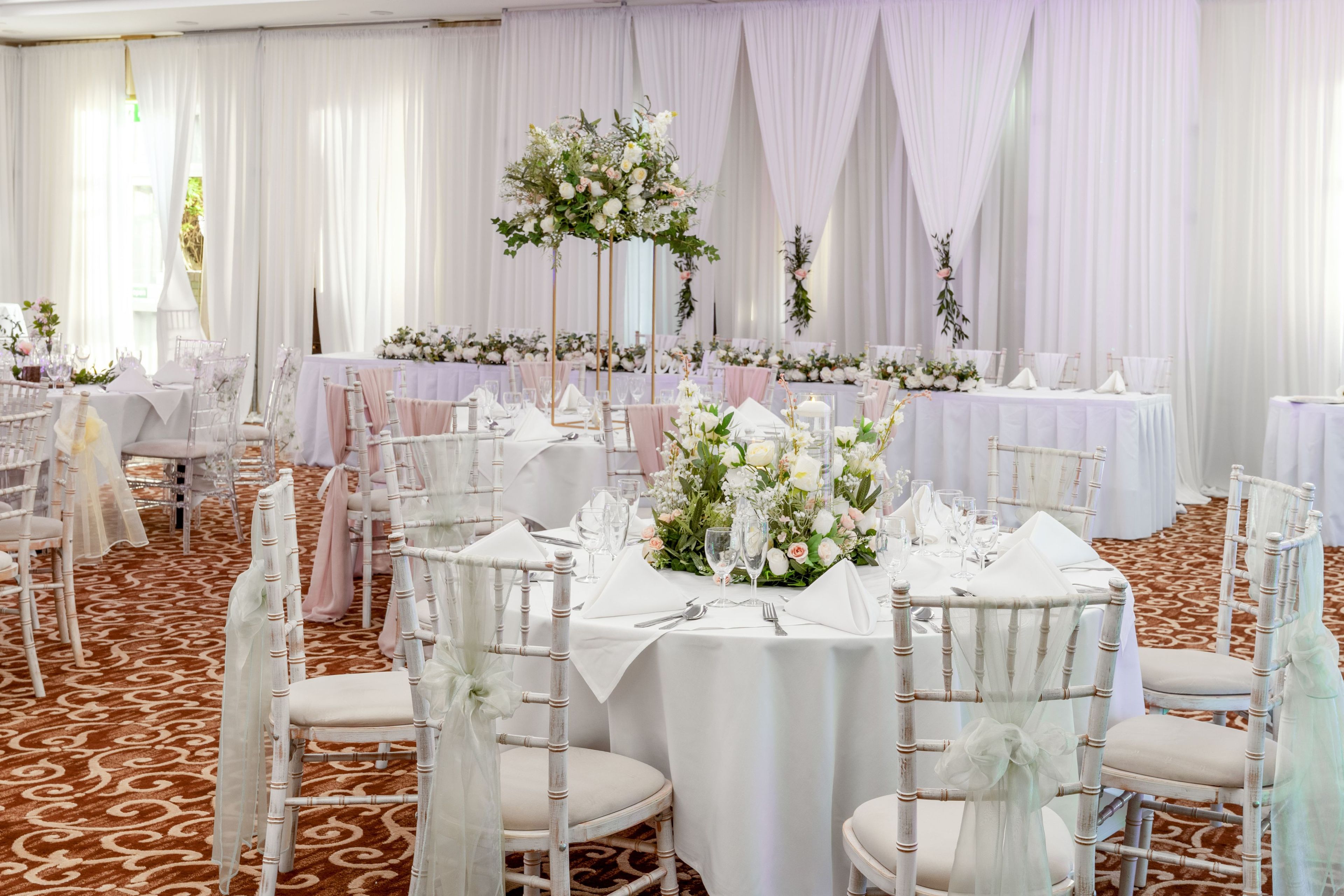 Un salón de banquetes con mesas redondas, sillas blancas y centros de mesa florales.