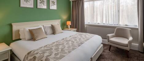 Een hotelkamer met een groot bed, een stoel, een nachtkastje, een lamp en een raam met gordijnen.
