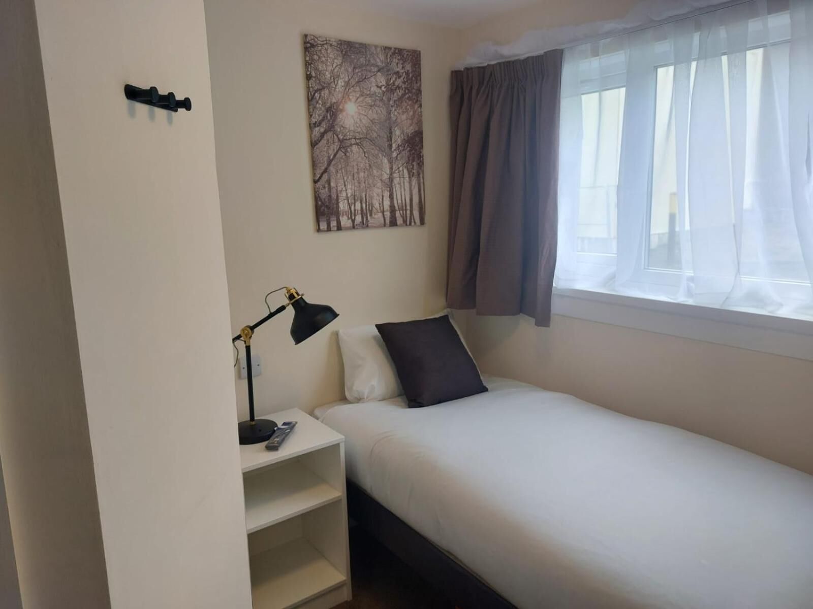 A small, single-bed room with a nightstand, a lamp, a remote control, and a painting on the wall.