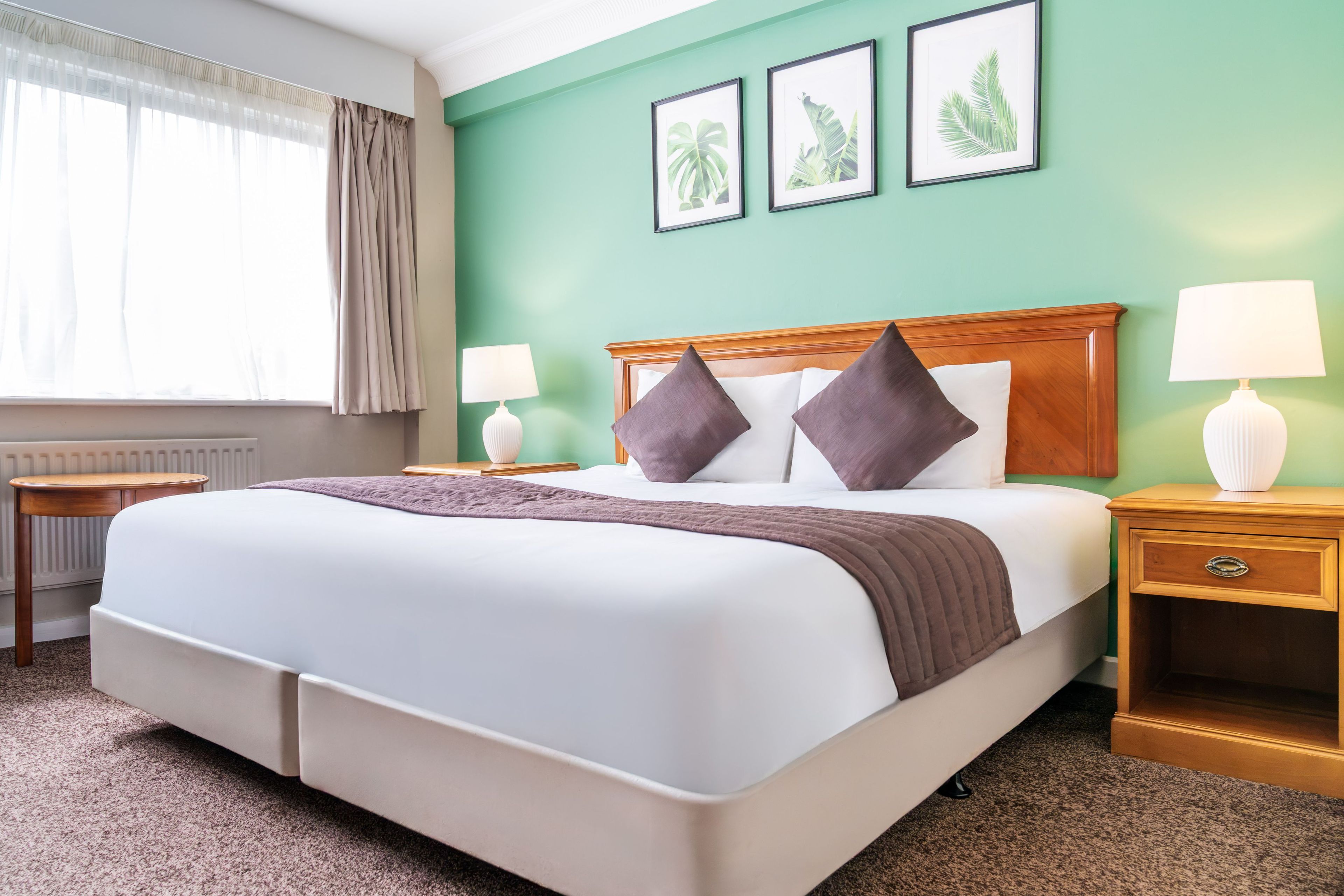 Habitación de hotel con una cama grande, dos mesitas de noche, un ventanal con cortinas y una pared verde con cuadros enmarcados.