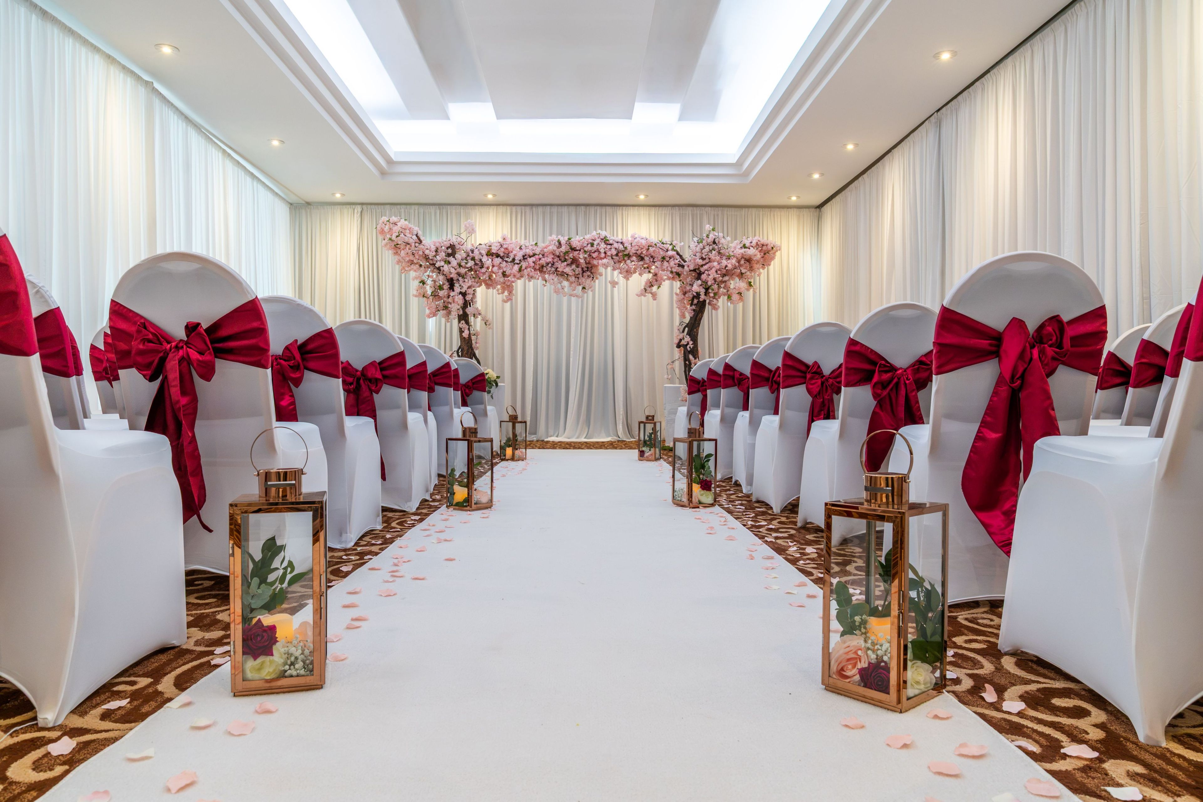 A wedding aisle with white chairs and red sashes, adorned with floral arrangements and lanterns.