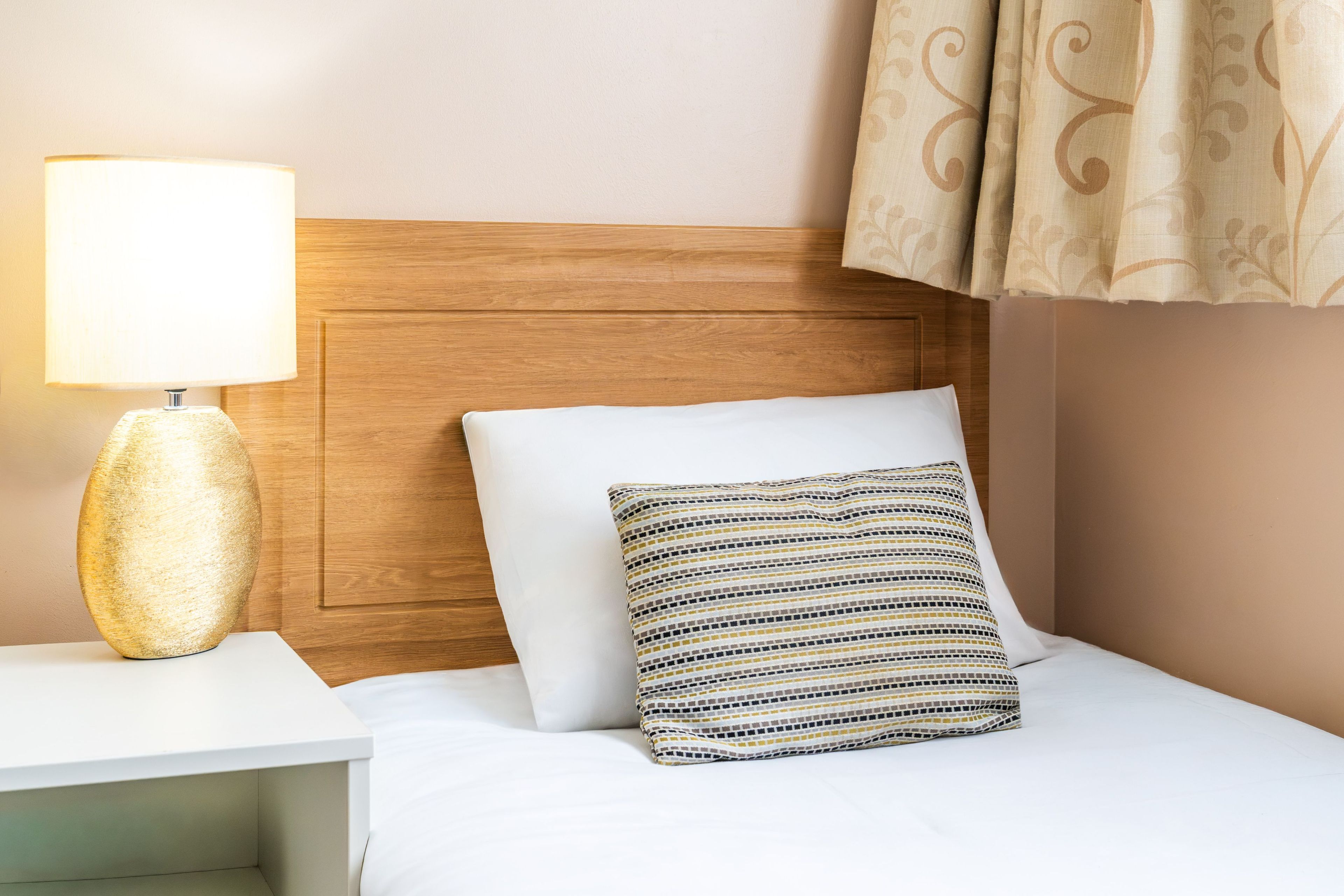 Habitación de hotel con cabecera de madera, mesita de noche blanca con lámpara, cama blanca con almohada estampada y cortinas color beige.