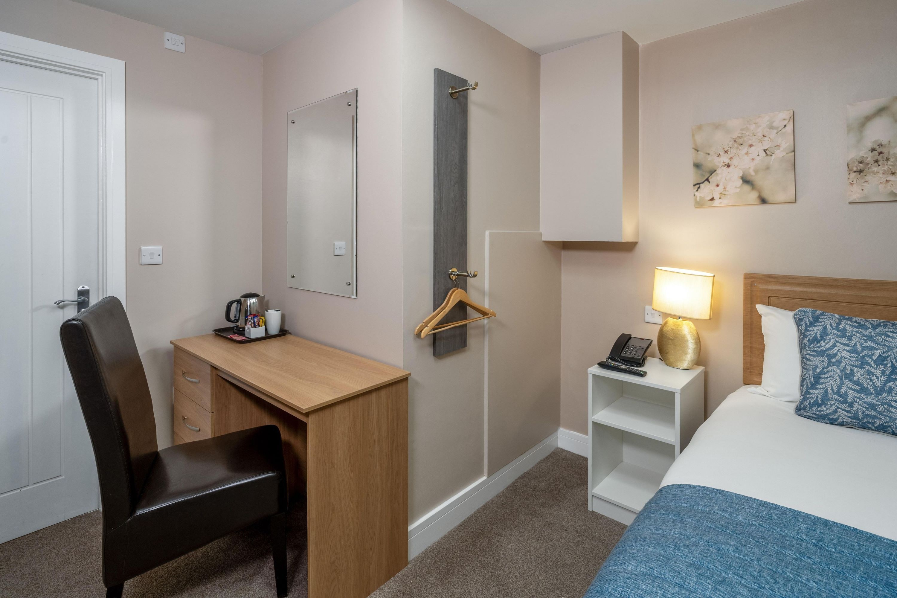 A hotel room with a desk, chair, bed, bedside table, lamp, and artwork on the wall.