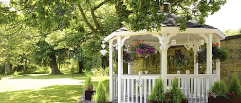 A gazebo with a white railing, surrounded by potted plants and hanging flower baskets.