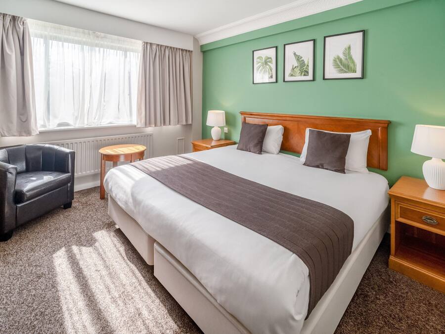 A hotel room with a large bed, a chair, a nightstand, a lamp, and a window with curtains.