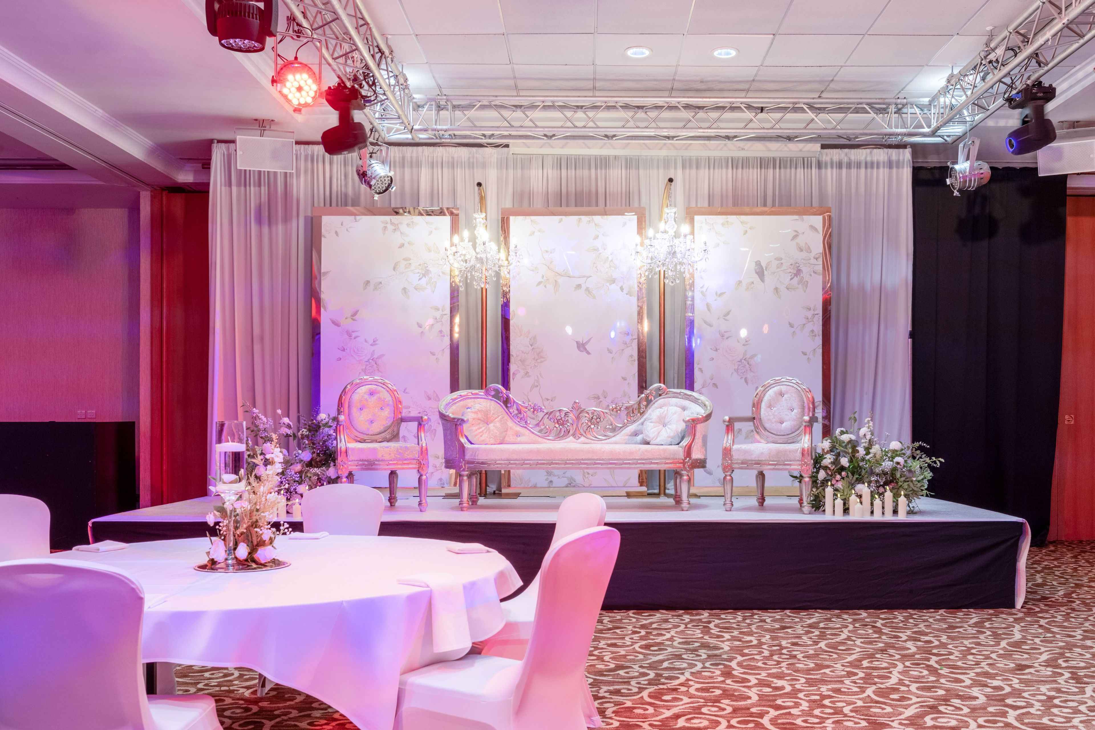 A stage with ornate chairs and a chandelier, surrounded by tables and chairs, in a room with patterned carpet and draped curtains.