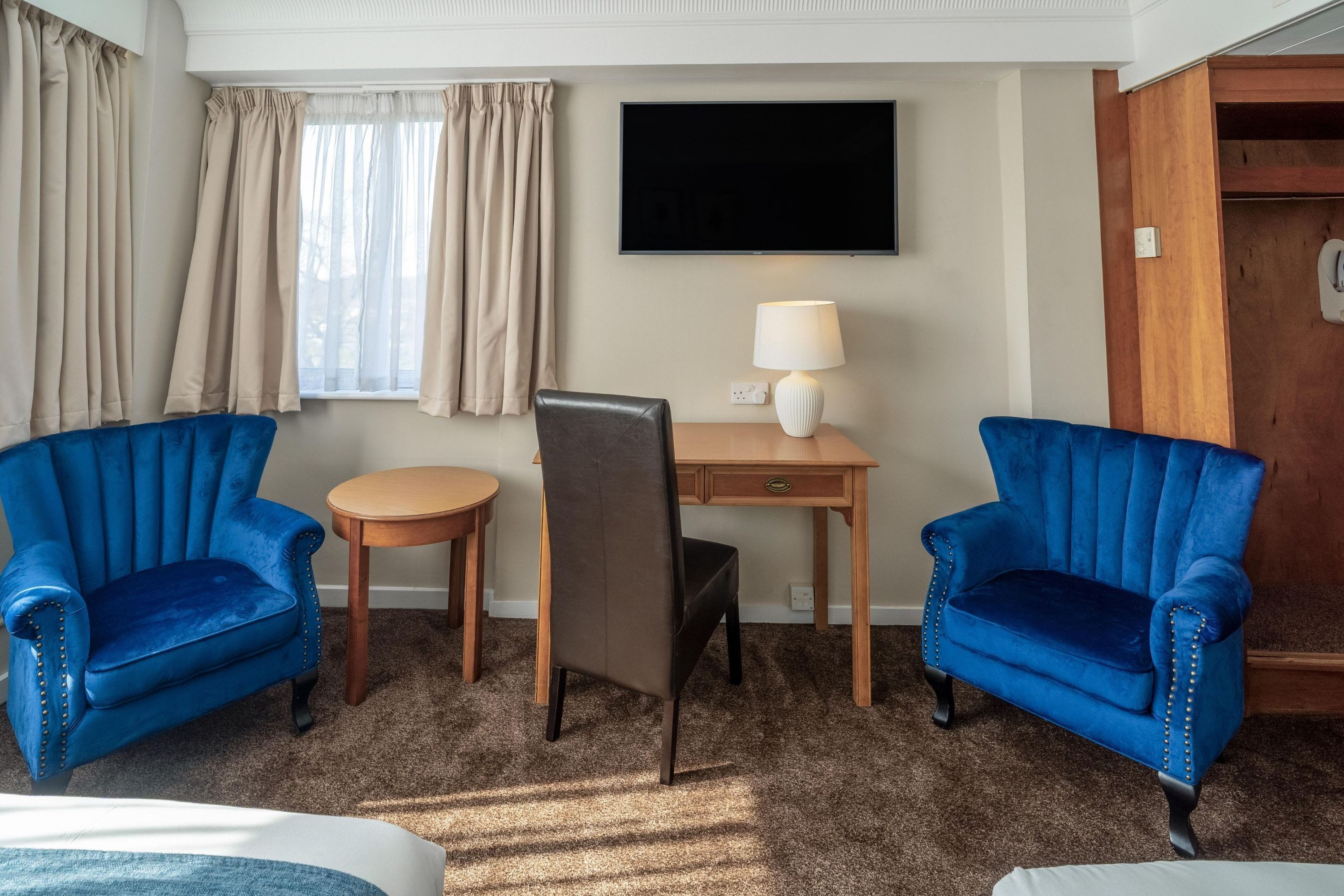 Een hotelkamer met twee blauwe fauteuils, een klein rond tafeltje, een bureau met een lamp, een televisie aan de muur en een kledingkast.