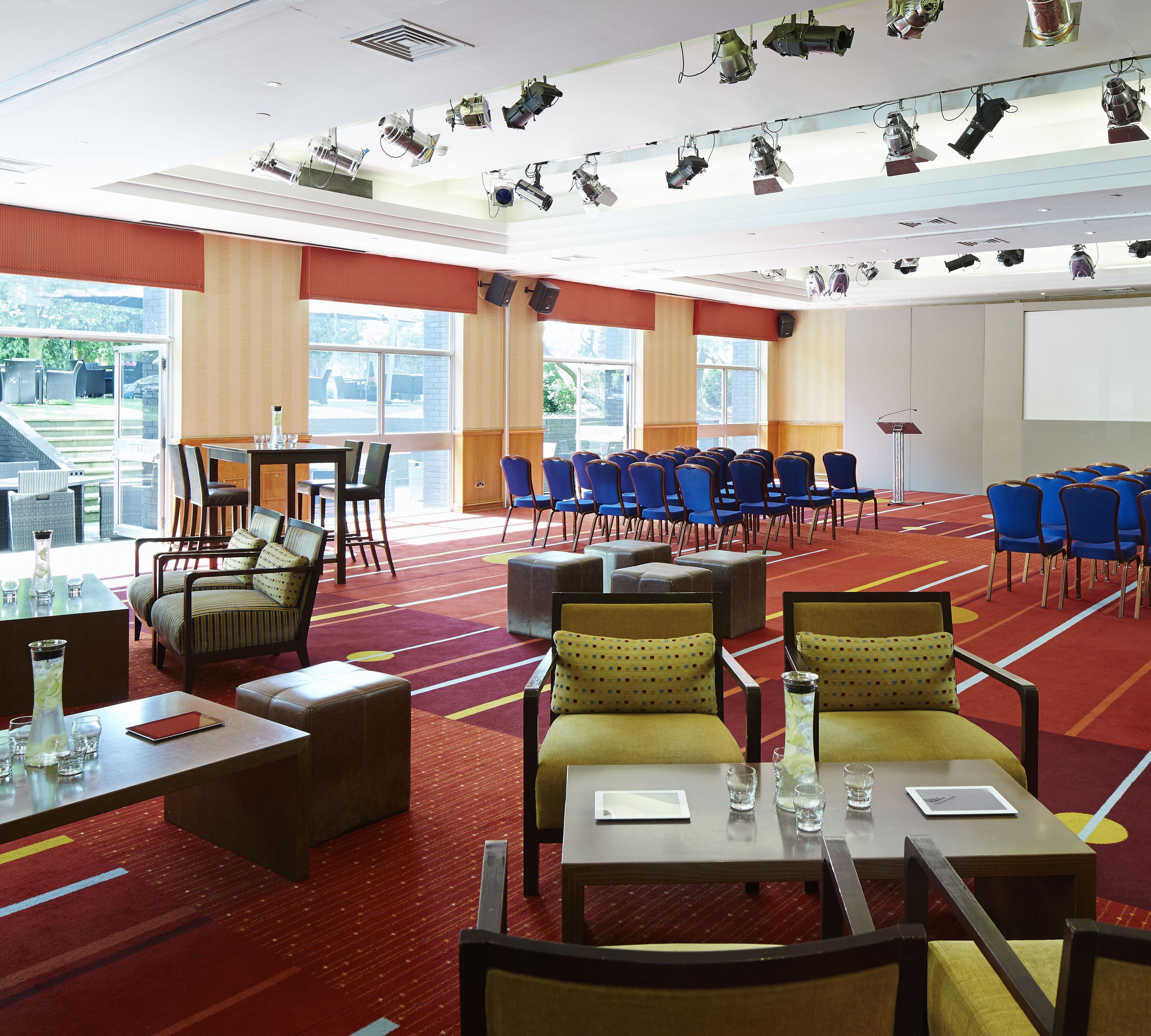 Een congreszaal met rijen blauwe stoelen, een spreekgestoelte en een scherm voor een beamer.
